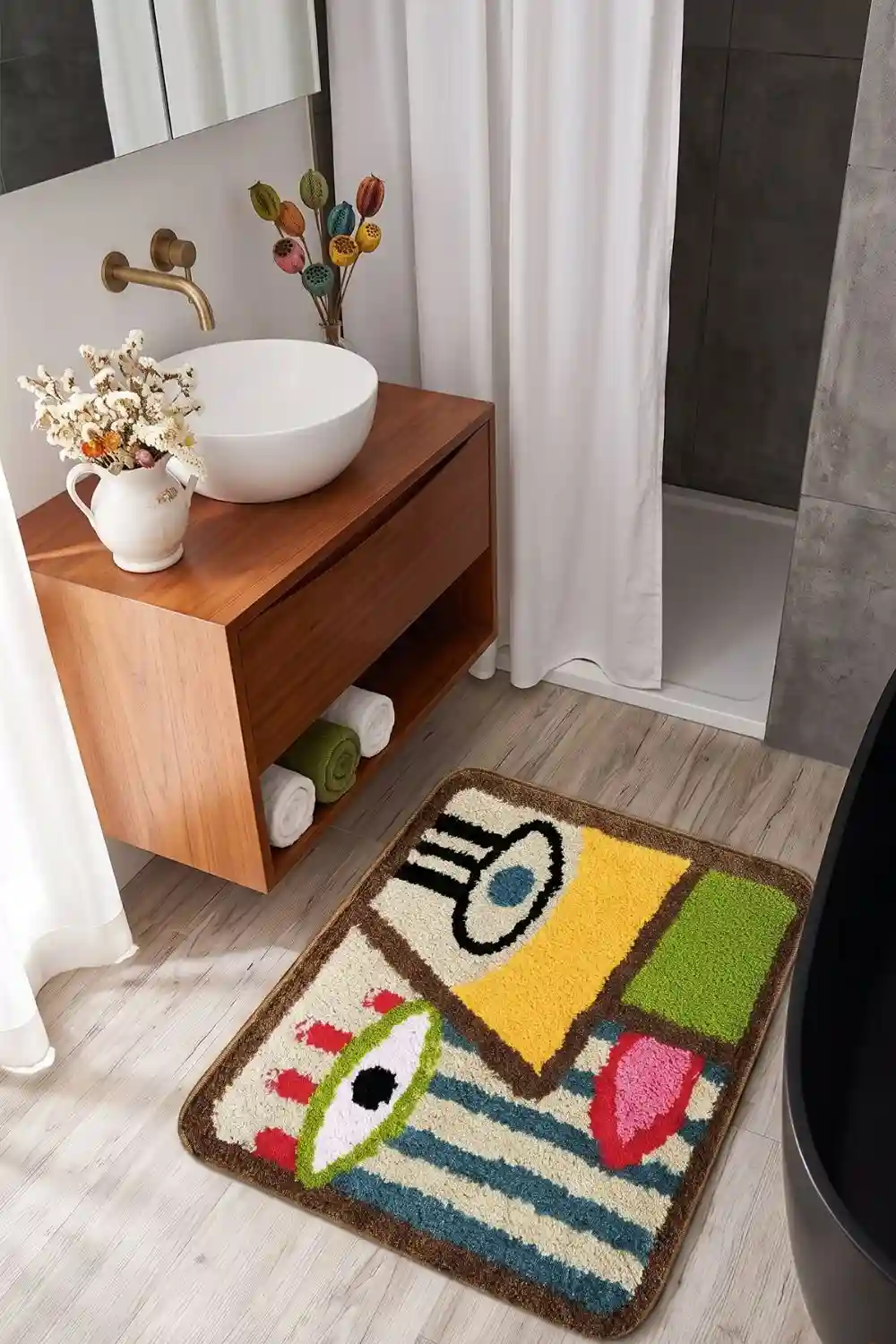 A modern bathroom scene featuring a small abstract eye-patterned bath mat on light wood flooring next to a floating vanity and black bathtub.